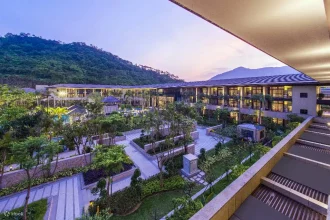 Modern resort near forested mountains with landscaped gardens, palm trees, and evening lights illuminating pathways.