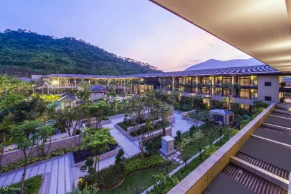 Modern resort near forested mountains with landscaped gardens, palm trees, and evening lights illuminating pathways.