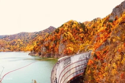 Large curved concrete dam near Sapporo beside turquoise reservoir with surrounding autumn forest in red, orange, and yellow.