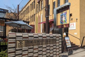 Entrance of the Kyoto International Manga Museum with a stone sign, yellow building, outdoor seating area, and museum poster display.