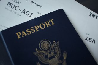 A close-up image of a U.S. passport resting on an international boarding pass, showing a partial view of flight details including gate, seat number, and flight route from Munich (MUC) to Ancona (AOI).