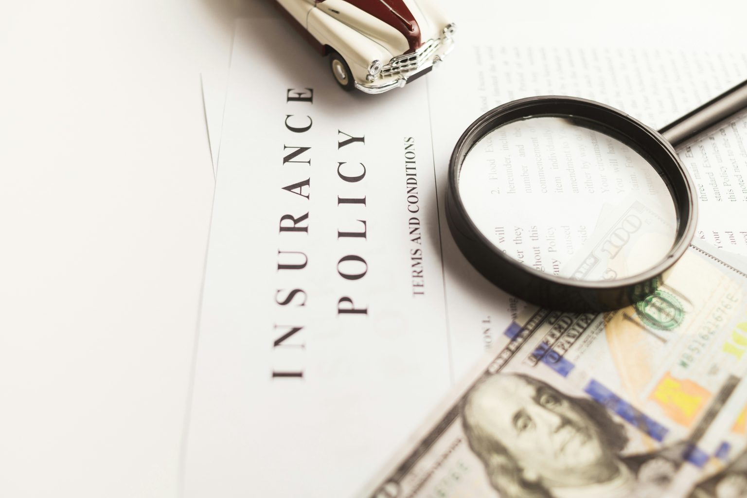 Close-up of an insurance policy document with a magnifying glass, a toy car, and U.S. hundred-dollar bills, symbolizing financial protection and vehicle coverage.