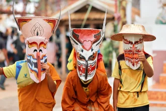 Three children wearing colorful Phi Ta Khon masks and traditional clothing during a festival in Thailand.