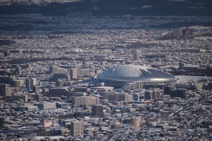 Snow-covered cityscape of Sapporo with a large silver dome stadium surrounded by buildings in winter.