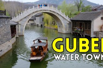 Wooden boat under stone arch bridge in Gubei Water Town with traditional houses and hills.