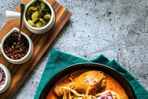 Bowl of khao soi curry noodles with chicken drumstick, crispy noodles, red onion, lime, and chili flakes.