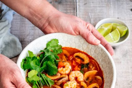 Hands holding a bowl of Thai Tom Yum soup with shrimp, mushrooms, cilantro, and lime in a spicy red broth.