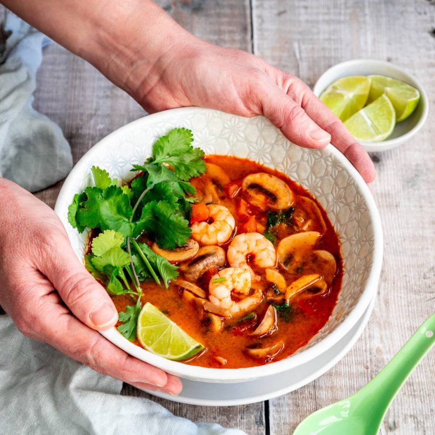 Hands holding a bowl of Thai Tom Yum soup with shrimp, mushrooms, cilantro, and lime in a spicy red broth.