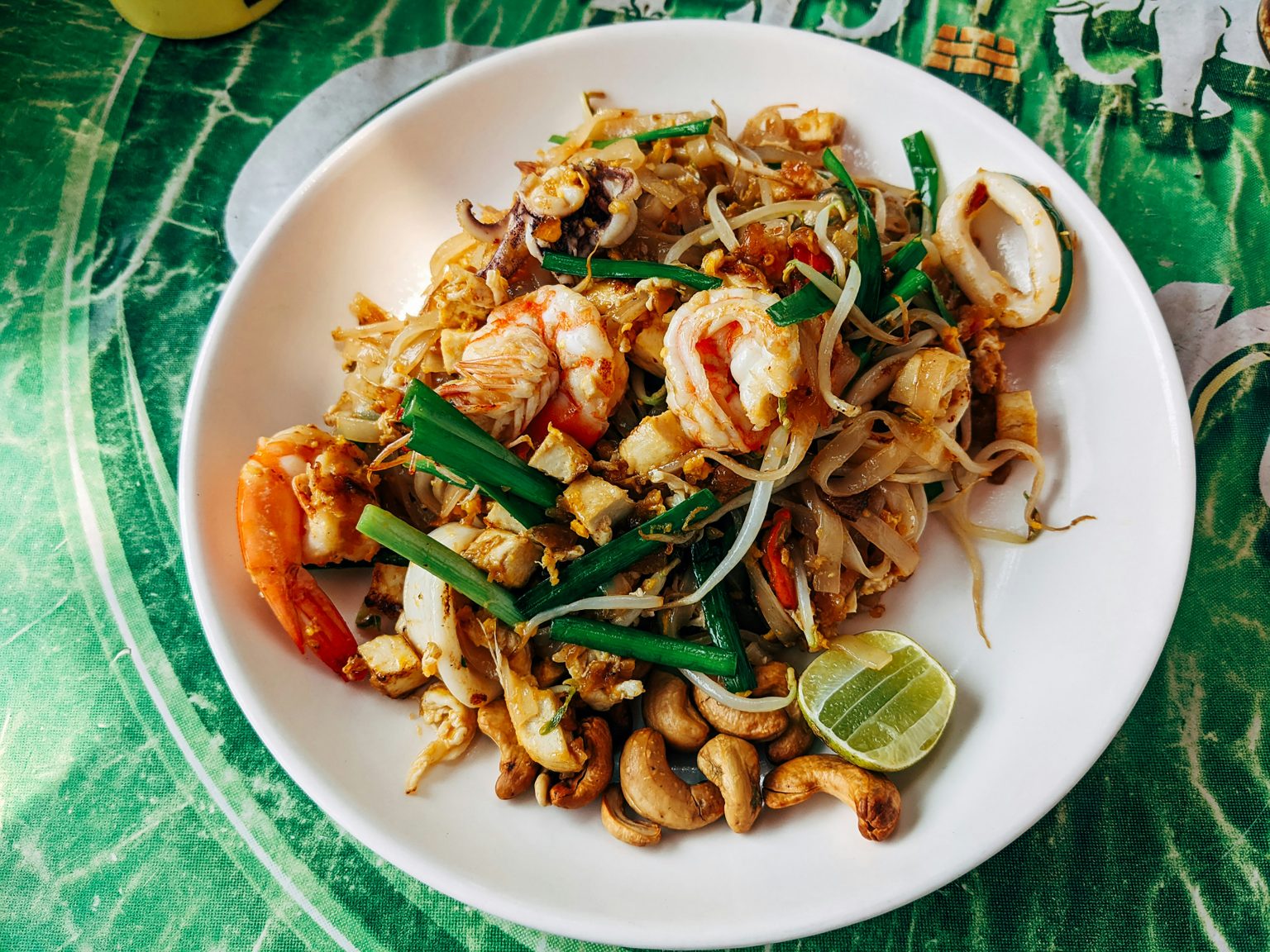 Plate of seafood Pad Thai topped with shrimp, squid, tofu, bean sprouts, green onions, and roasted cashews, garnished with a lime wedge.